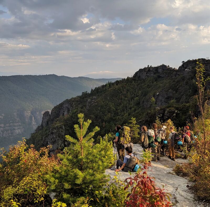 A group of hikers is walking along a narrow, rocky trail on a mountain ridge. The trail is surrounded by lush green vegetation and trees. In the distance, there are more mountains and a cloudy sky. The hikers appear to be enjoying the scenic views and the natural beauty of their surroundings.